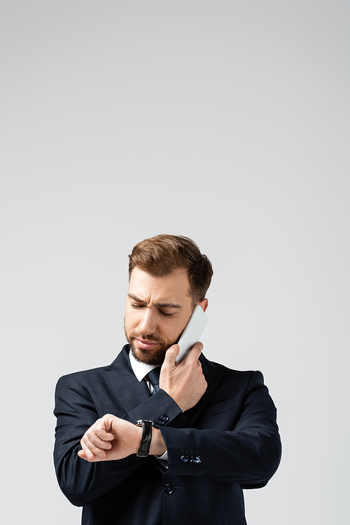 serious handsome businessman in suit talking on smartphone and looking at wristwatch isolated on grey