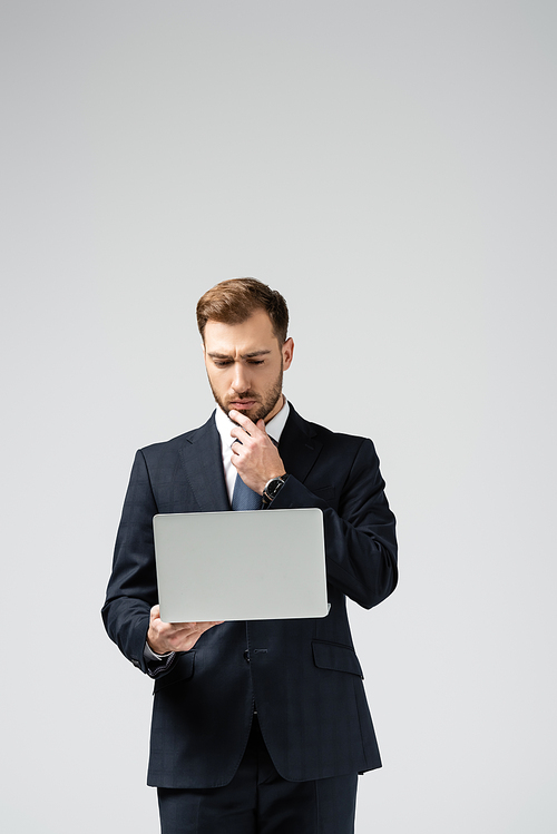 pensive handsome businessman in suit using laptop isolated on grey