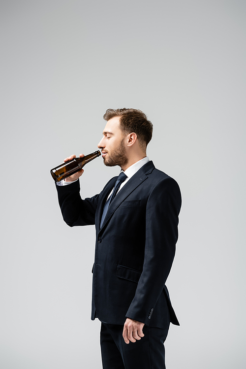 side view of handsome businessman in suit drinking beer isolated on grey
