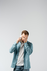 confused young man talking on smartphone and showing facepalm isolated on grey