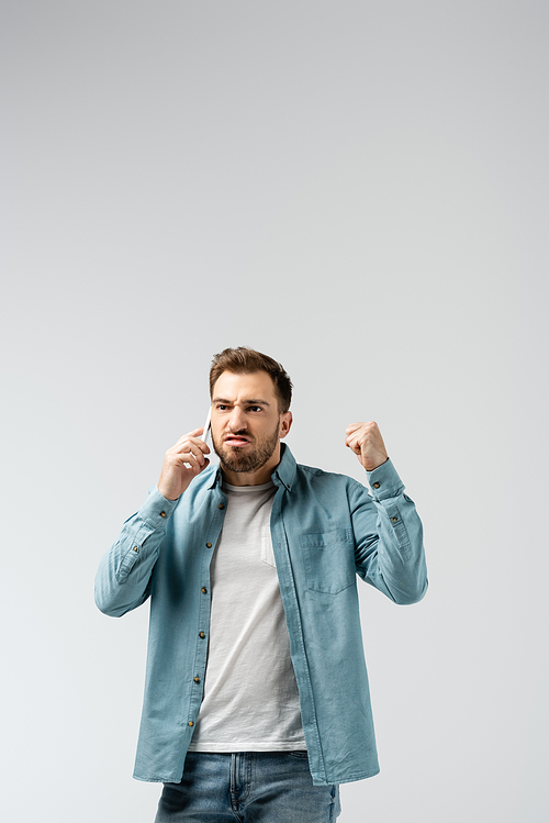 angry young man talking on smartphone and showing fist isolated on grey