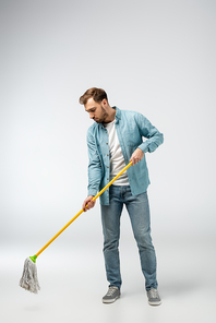 sad young man cleaning floor with mop isolated on grey