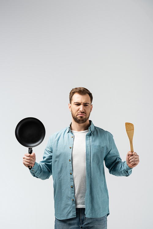 displeased young man with frying pan and spatula isolated on grey