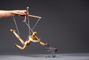 cropped view of puppeteer holding wooden marionette with shopping cart on black