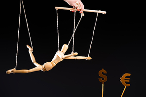 cropped view of puppeteer holding wooden marionette on strings near currency signs isolated on black
