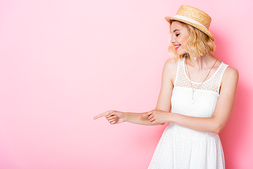 woman in white dress and straw hat pointing with fingers and looking away on pink