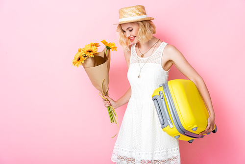 young woman in straw hat holding yellow flowers and luggage on pink