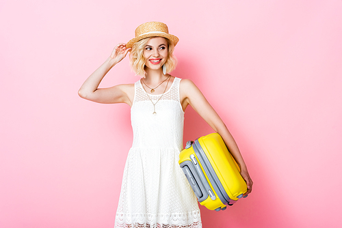 young woman touching straw hat while holding yellow luggage on pink