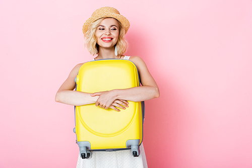 woman in straw hat hugging yellow luggage on pink