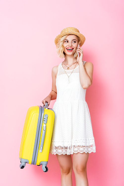 woman in straw hat holding luggage and talking on smartphone on pink