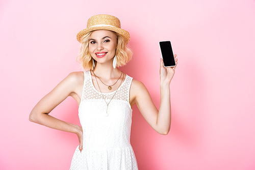 woman in straw hat holding smartphone with blank screen and standing with hand on hip on pink