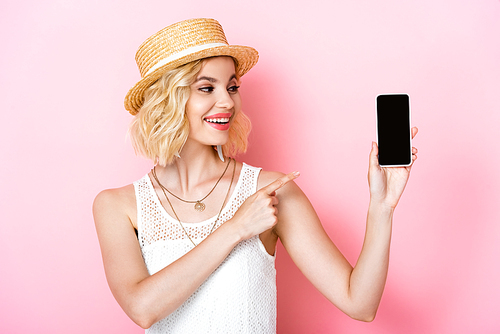 woman in straw hat pointing with finger at smartphone with blank screen on pink