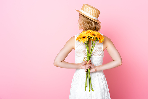 back view of young woman in straw hat and white dress holding yellow flowers behind back on pink