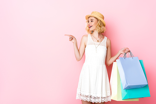 woman in straw hat pointing with finger and holding shopping bags on pink
