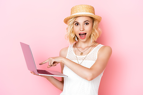 excited woman in straw hat pointing with finger at laptop on pink