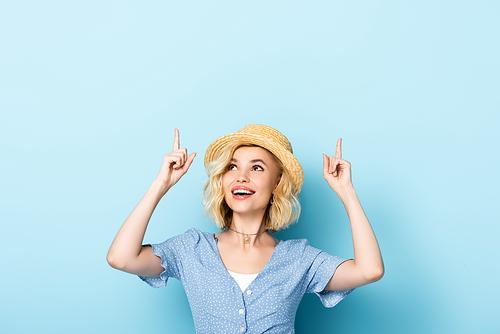 young woman in straw hat pointing with fingers and looking up on blue