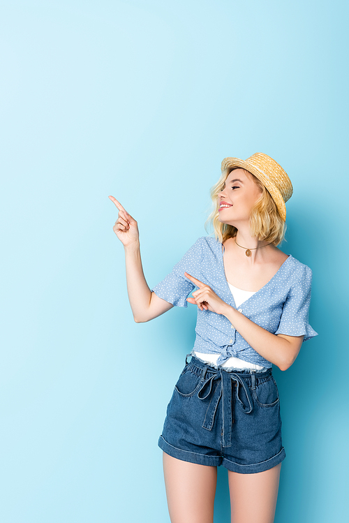 woman in straw hat pointing with fingers and looking away on blue
