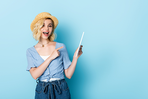 excited woman in straw hat and shorts pointing with finger at digital tablet on blue