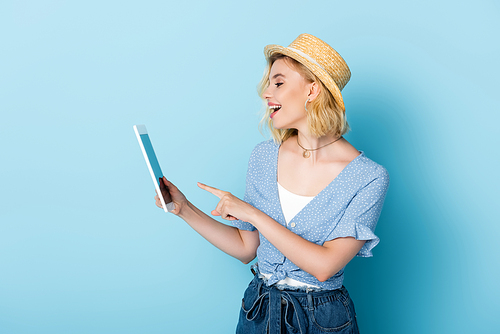 excited woman in straw hat pointing with finger at digital tablet on blue