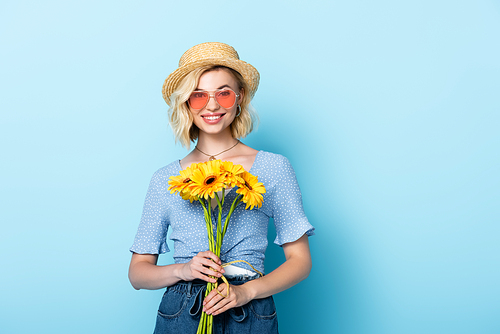 young woman in straw hat and sunglasses holding yellow flowers while standing on blue