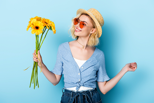 young woman in straw hat and sunglasses holding flowers while standing on blue