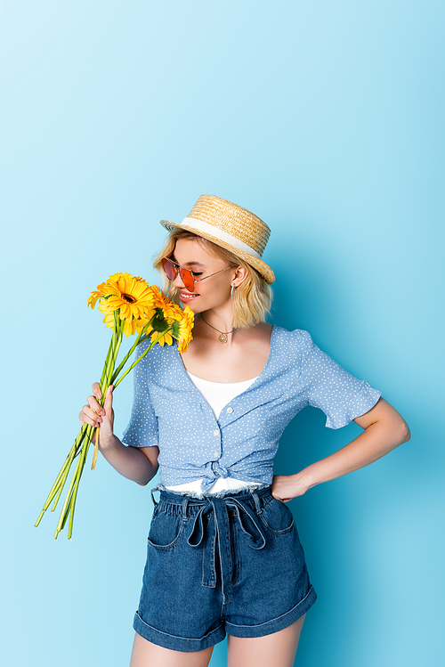young woman in straw hat and sunglasses smelling yellow flowers while standing with hand on hip on blue