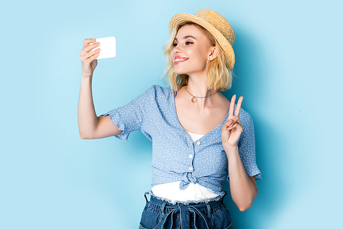 young woman in straw hat showing peace sign and taking selfie on blue