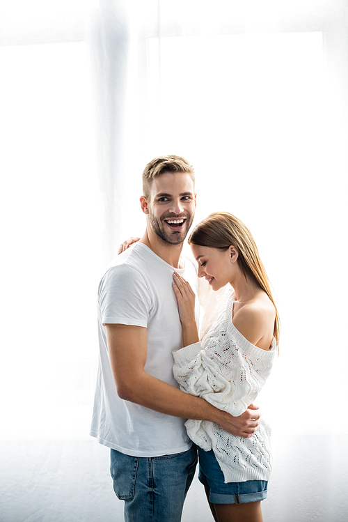handsome man hugging attractive and smiling woman in apartment