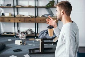 side view of shocked man dropping paper cup and looking at robbed apartment