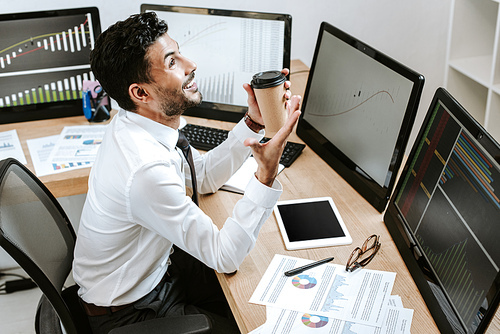 high angle view of smiling bi-racial trader holding paper cup and sitting near computers with graphs