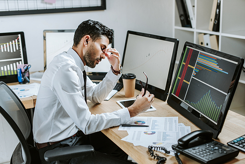 side view of tired bi-racial trader sitting near computers with graphs and holding glasses