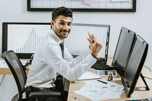 smiling bi-racial trader showing thumbs up and sitting near computers with graphs