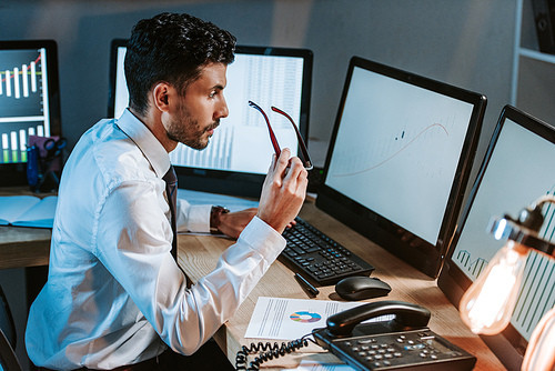 side view of bi-racial trader holding glasses and looking at computer with graphs