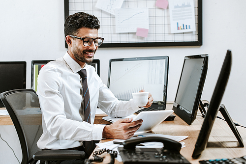 smiling bi-racial trader using digital tablet and holding cup