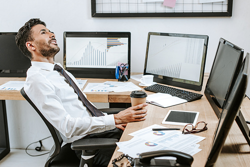 smiling bi-racial trader holding paper cup and sitting near computers with graphs