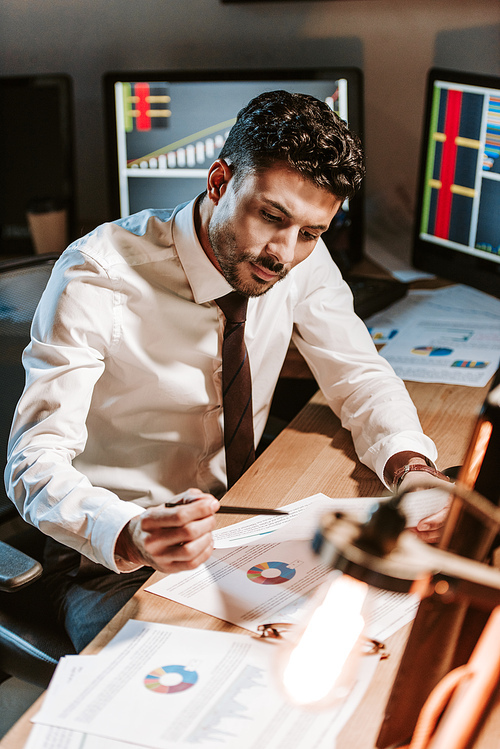 bi-racial trader looking at paper and sitting near computers with graphs