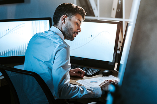 bi-racial trader looking at paper and sitting near computers with graphs