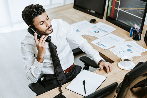 high angle view of bi-racial trader talking on telephone and sitting near computer with graphs