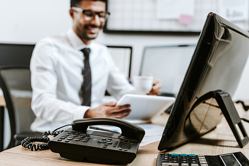 selective focus of telephone and calculator on table and smiling trader with digital tablet on background