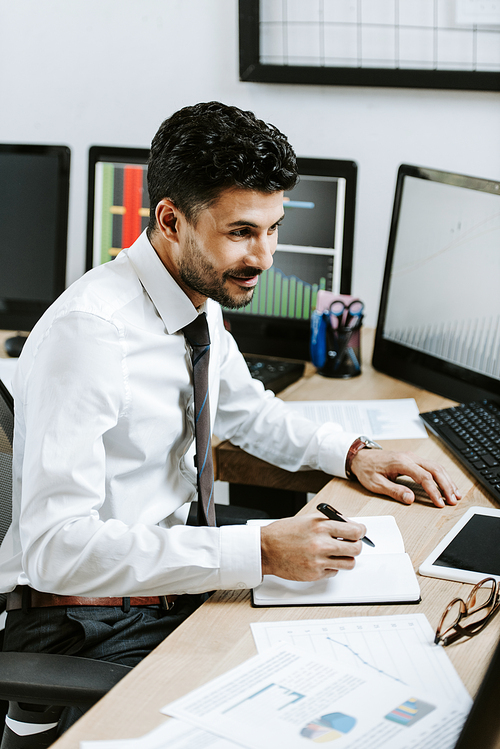 smiling bi-racial trader writing and sitting near computers with graphs