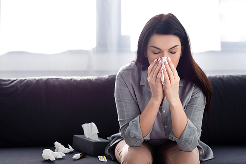 woman with allergy sneezing in napkin while sitting on sofa