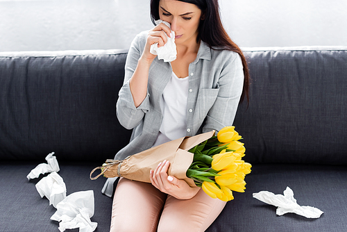sneezing woman with pollen allergy sitting on sofa with flowers