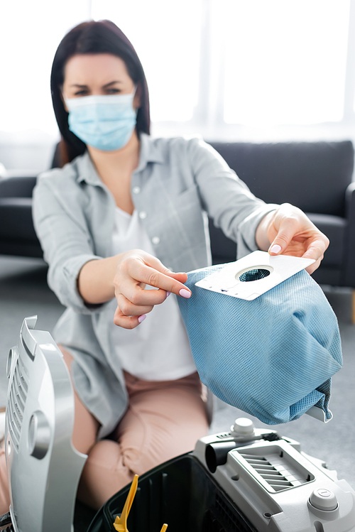 selective focus of allergic woman in medical mask holding dust bag near vacuum cleaner in living room