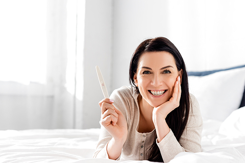 cheerful and pregnant woman holding pregnancy test and lying on bed