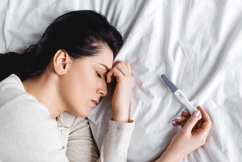 top view of depressed woman with closed eyes lying on bed near pregnancy test