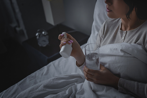 cropped view of woman with sleep disorder holding glass of water and bottle with sleeping pills in bedroom