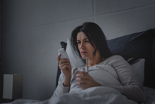woman with sleep disorder holding glass of water and bottle with sleeping pills in bedroom