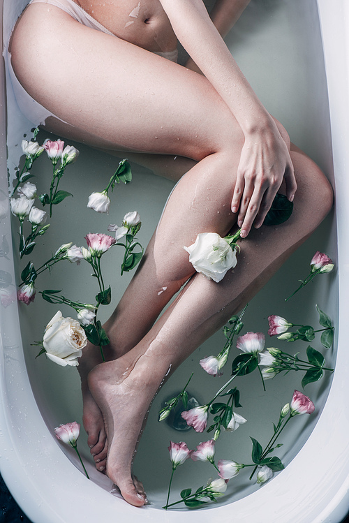 top view of beautiful female legs in clear water with flowers in white bathtub