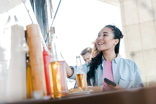 selective focus of happy asian girl smiling near bottles with beer and customers near food truck