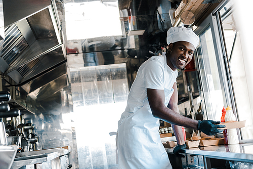 selective focus of cheerful african american chef smiling while holding carton plate in food truck